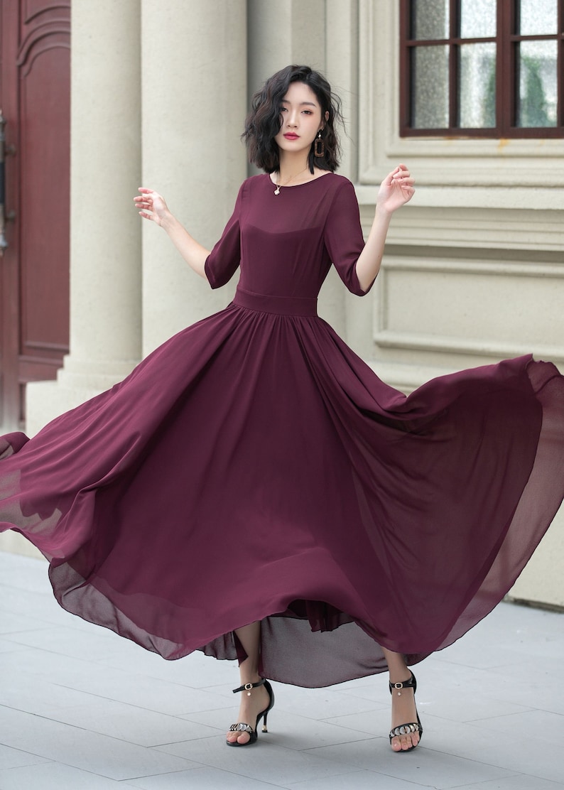 May include: A woman wearing a long, flowing burgundy dress with a round neckline and 3/4 sleeves. The dress has a fitted bodice and a full skirt. She is wearing black heeled sandals with rhinestones.
