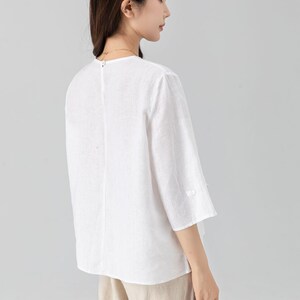 May include: White linen top with three-quarter sleeves and a round neckline. The blouse has a relaxed fit and a button detail on the sleeves. The model is wearing beige pants. This top is suitable for casual wear.