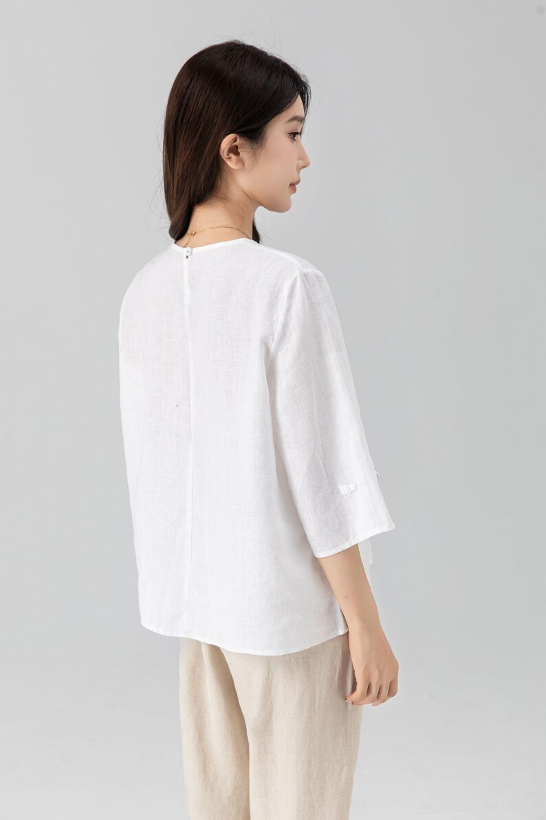May include: White linen top with three-quarter sleeves and a round neckline. The blouse has a relaxed fit and a button detail on the sleeves. The model is wearing beige pants. This top is suitable for casual wear.