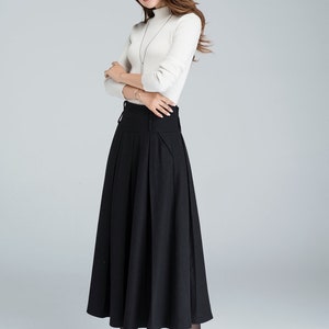 May include: A black high-waisted skirt with pleats and a wide waistband. The skirt is midi-length and falls to the calves. The model is wearing a white turtleneck sweater and black heels.