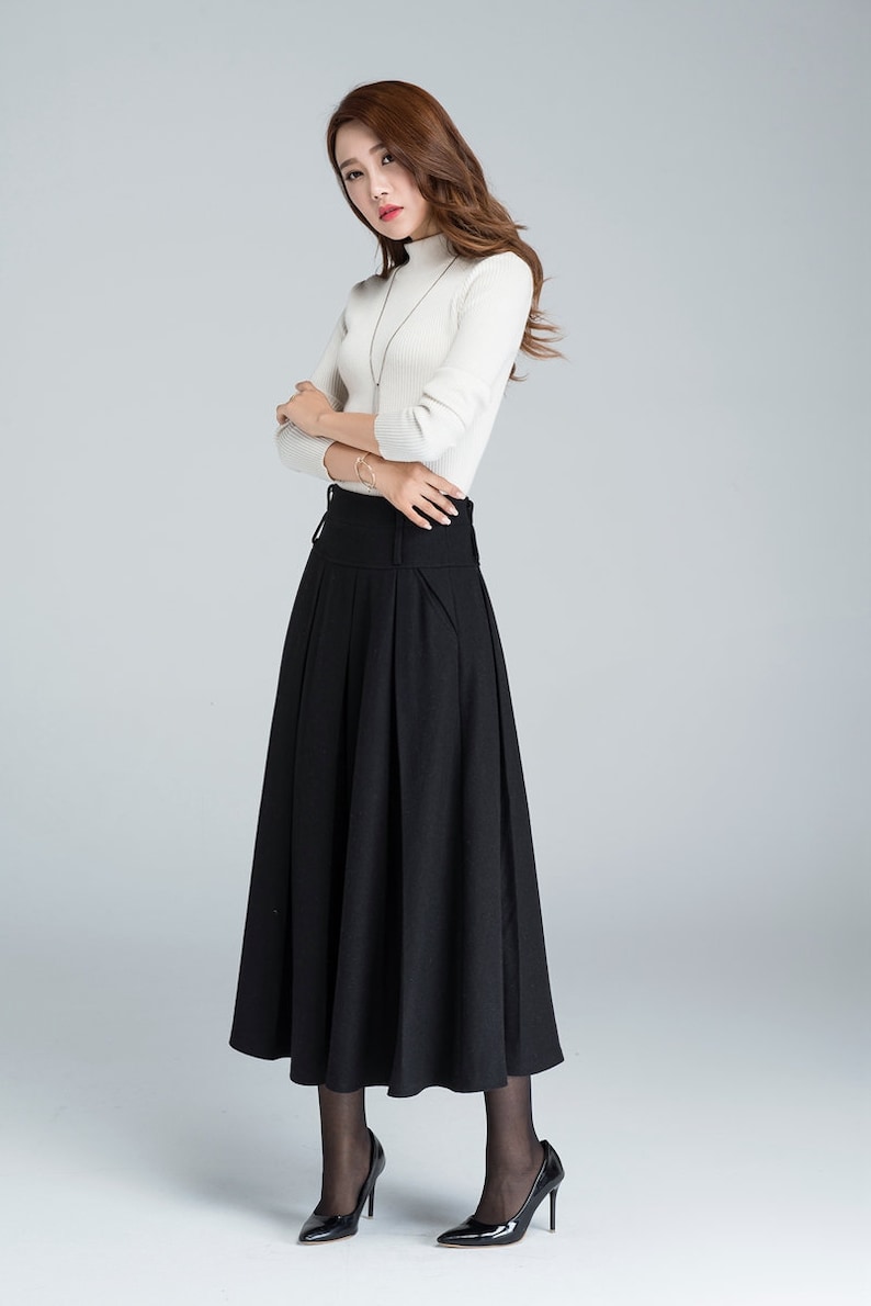 May include: A black high-waisted skirt with pleats and a wide waistband. The skirt is midi-length and falls to the calves. The model is wearing a white turtleneck sweater and black heels.
