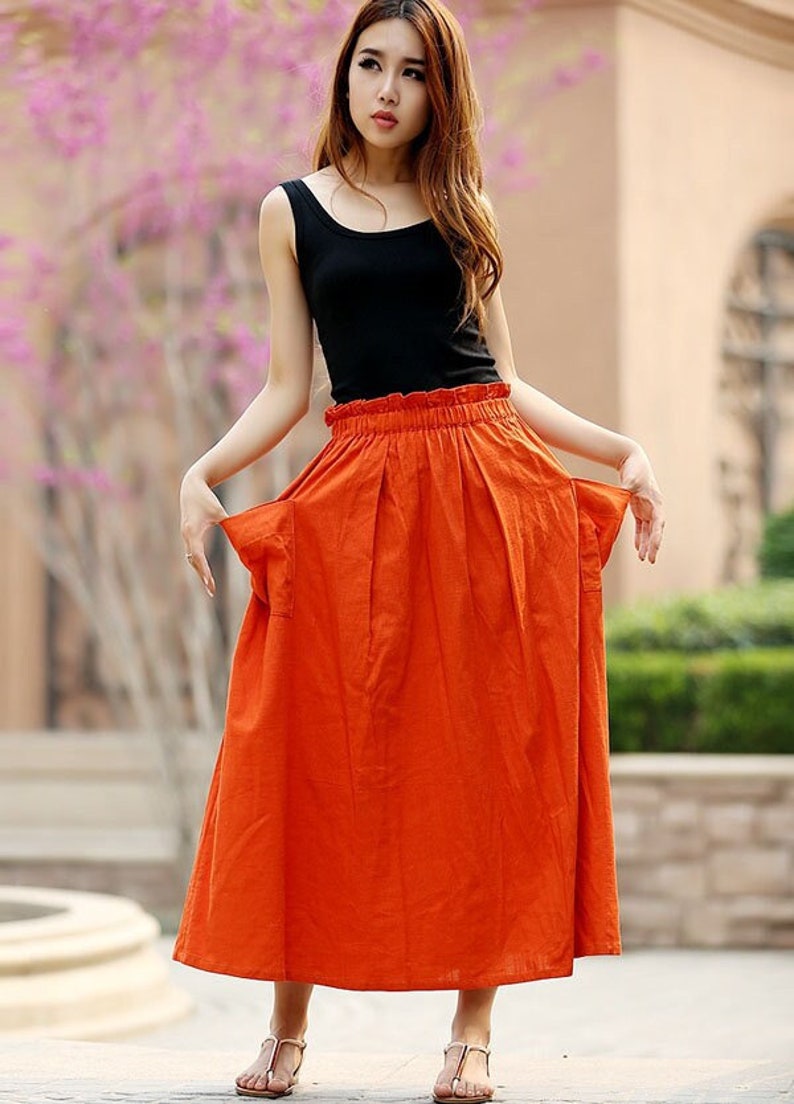 May include: A black tank top paired with a long, vibrant orange skirt. The skirt has a high waist and a flowing, pleated design. The outfit is completed with simple sandals, creating a casual yet stylish look.