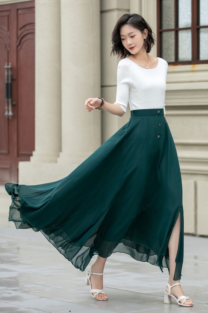 May include: A woman wearing a white short-sleeved top and a long, dark green skirt with a front slit and button detailing. The skirt is flowing and the woman is walking.