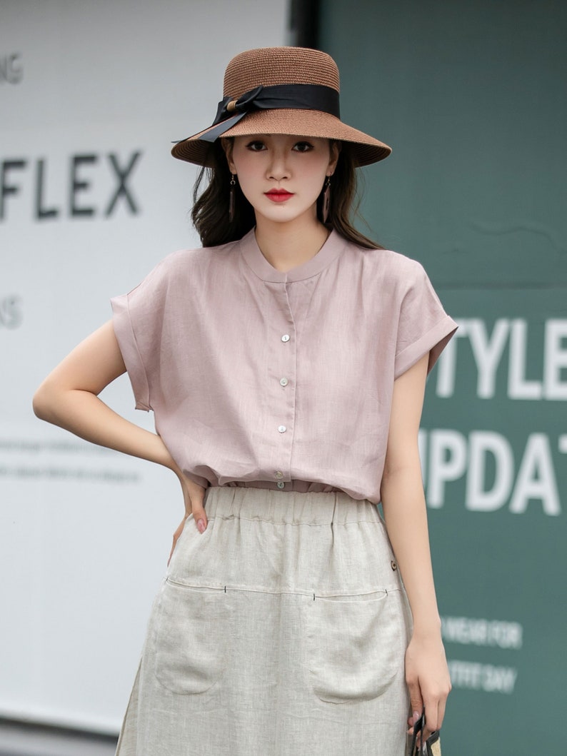 May include: A woman wearing a light pink linen blouse with a button-up front and short sleeves. She is also wearing a beige linen skirt with two large pockets on the front. She is wearing a brown straw hat with a black ribbon.