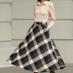 May include: A woman wearing a beige plaid button-down shirt and a long, flared plaid skirt in shades of brown, black, and cream. The outfit is paired with dark brown ankle boots. The skirt has pockets.