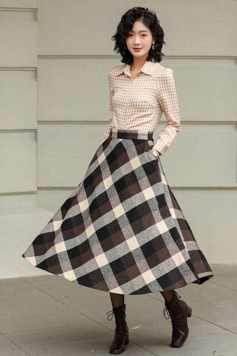 May include: A woman wearing a beige plaid button-down shirt and a long, flared plaid skirt in shades of brown, black, and cream. The outfit is paired with dark brown ankle boots. The skirt has pockets.