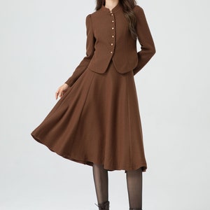 May include: A brown skirt suit with a fitted jacket and a flared skirt. The jacket has a mandarin collar and gold buttons. The model is wearing black tights and brown lace-up boots.