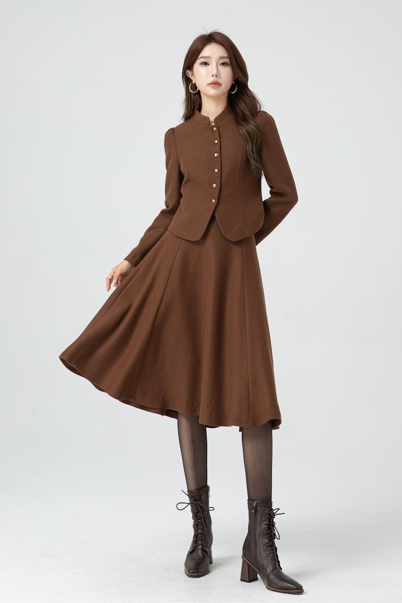 May include: A brown skirt suit with a fitted jacket and a flared skirt. The jacket has a mandarin collar and gold buttons. The model is wearing black tights and brown lace-up boots.