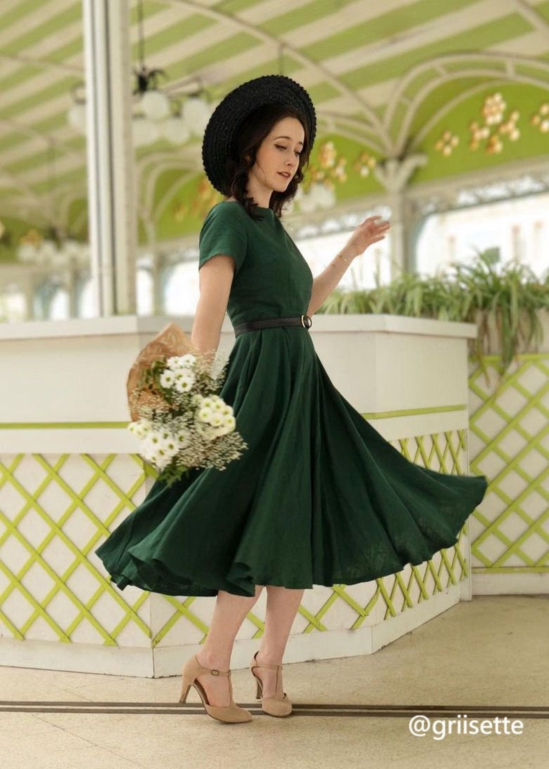 May include: A woman wearing a dark green dress with a black belt and a black hat, holding a bouquet of white flowers. She is standing in a green and white tiled area.