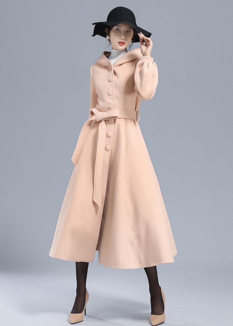 May include: A long, light pink wool coat with a hood, a belt, and button closures. The coat has a flared skirt and long sleeves.