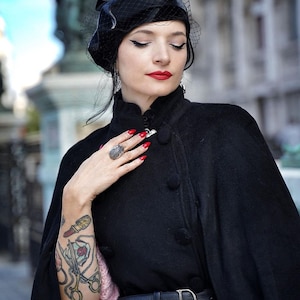 May include: A woman wearing a black cape with a black beret and a black veil. She has red lipstick and red nail polish. She is wearing a silver ring with a black stone on her ring finger. She has a tattoo on her arm of a lipstick, scissors, and a rose.