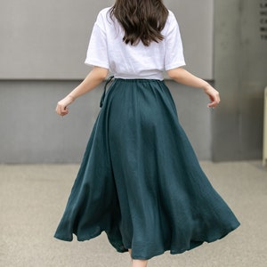 May include: A woman wearing a white t-shirt and a dark green maxi skirt with a brown belt. She is wearing a straw hat with a brown band and brown sandals.
