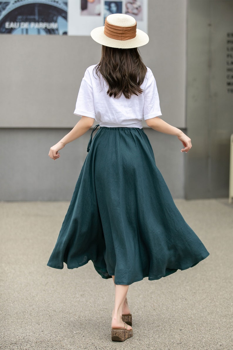 May include: A woman wearing a white t-shirt and a dark green maxi skirt with a brown belt. She is wearing a straw hat with a brown band and brown sandals.