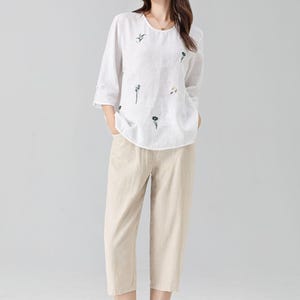 May include: A white linen top with embroidered floral details and three-quarter sleeves paired with beige cropped pants. The outfit is a casual, comfortable style, suitable for everyday wear. The ensemble is a light, airy design.