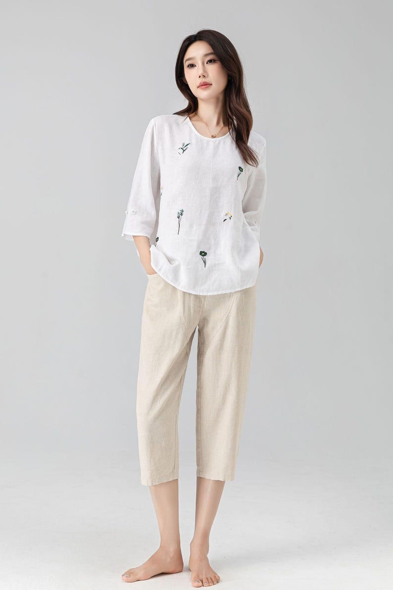 May include: A white linen top with embroidered floral details and three-quarter sleeves paired with beige cropped pants. The outfit is a casual, comfortable style, suitable for everyday wear. The ensemble is a light, airy design.