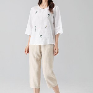 May include: A white linen top with embroidered floral details and three-quarter sleeves is paired with beige capri pants. The outfit is a casual, comfortable style, suitable for warm weather. The ensemble is a relaxed fit.