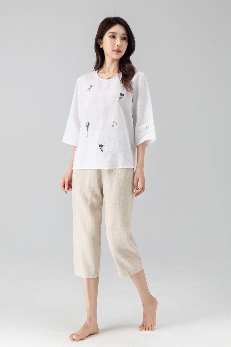 May include: A white linen top with embroidered floral details and three-quarter sleeves is paired with beige capri pants. The outfit is a casual, comfortable style, suitable for warm weather. The ensemble is a relaxed fit.