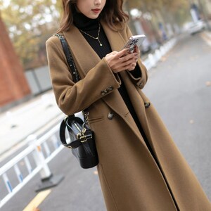 May include: A brown double-breasted long coat with a black turtleneck sweater underneath. The person is holding a smartphone in their right hand.
