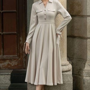 May include: A beige long-sleeved dress with a collared neckline, button closure, and a fitted bodice. The dress has a flared skirt and tie details at the cuffs.