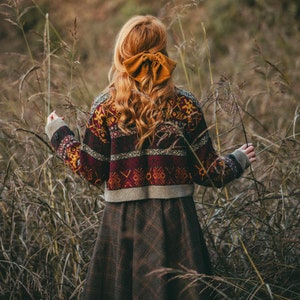 Pu&ograve; includere: Una donna indossa un maglione a motivi marroni e rossi con una gonna a quadri marroni e beige. Ha i capelli lunghi e ricci rossi con un grande fiocco giallo nei capelli. &Egrave; in piedi in un campo di erba alta.