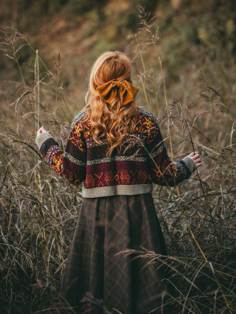 Pu&ograve; includere: Una donna indossa un maglione a motivi marroni e rossi con una gonna a quadri marroni e beige. Ha i capelli lunghi e ricci rossi con un grande fiocco giallo nei capelli. &Egrave; in piedi in un campo di erba alta.