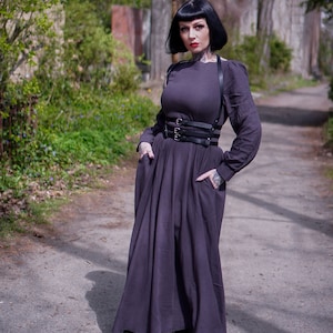 May include: A woman wearing a long, dark grey dress with a black leather harness and a black belt. She is standing on a path in a park.