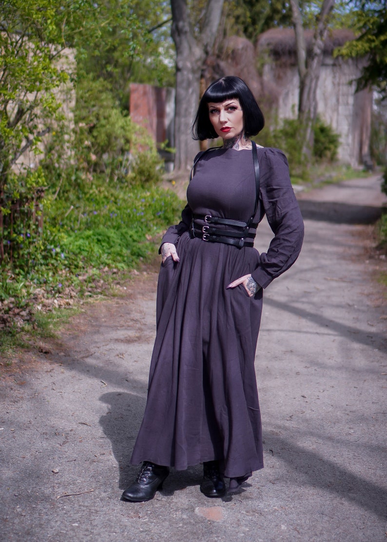 May include: A woman wearing a long, dark grey dress with a black leather harness and a black belt. She is standing on a path in a park.
