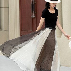 May include: A woman wearing a black top and a white hat is walking in a long, flowing skirt with black and white panels. The skirt is sheer and has a ruffled hem.