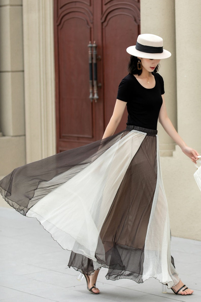 May include: A woman wearing a black top and a white hat is walking in a long, flowing skirt with black and white panels. The skirt is sheer and has a ruffled hem.