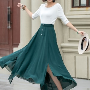 May include: A woman wearing a white top and a long, dark green skirt with a front slit. The skirt has a button closure and is flowing in the wind. She is wearing white sandals and holding a straw hat.