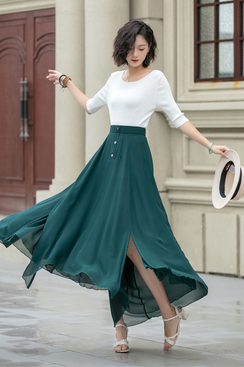 May include: A woman wearing a white top and a long, dark green skirt with a front slit. The skirt has a button closure and is flowing in the wind. She is wearing white sandals and holding a straw hat.
