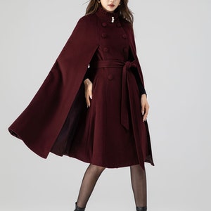 Pu&ograve; includere: Un cappotto a mantella color bordeaux scuro in misto lana con collo alto a volant, bottoni effetto perla, chiusura doppiopetto e cintura in tessuto abbinata. Il capo presenta una silhouette svasata con aperture per le braccia.