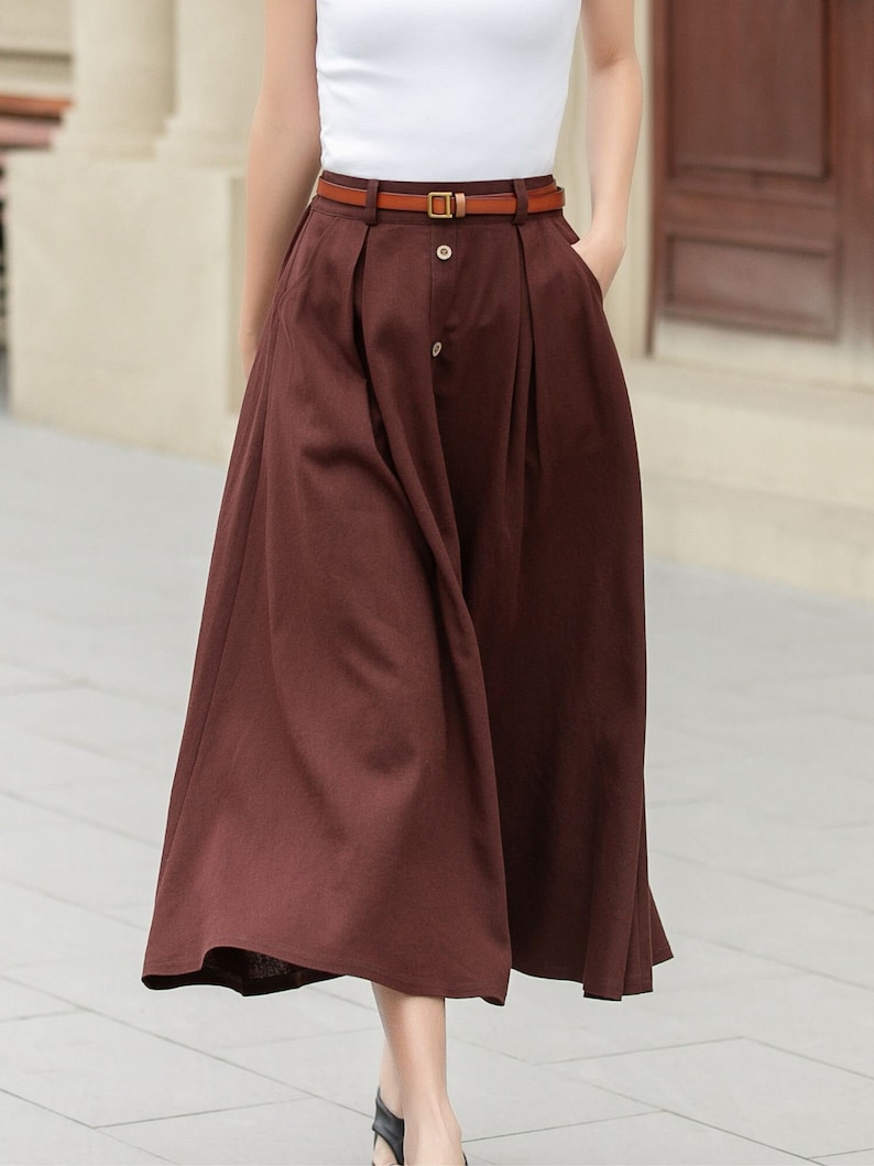 May include: A brown midi skirt with a brown leather belt and a gold buckle. The skirt has a button closure and pleats.