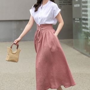 May include: A woman wearing a white short-sleeved shirt with a button-up front and a pink linen skirt with a lace-up detail at the waist. She is carrying a small straw bag and wearing silver heeled sandals.