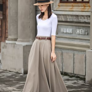 May include: A woman wearing a white top with elbow-length sleeves, a long beige skirt, and a brown belt. She is also wearing a brown hat with a black band and black high heels. The background features a building with the text "HUAN HENG".