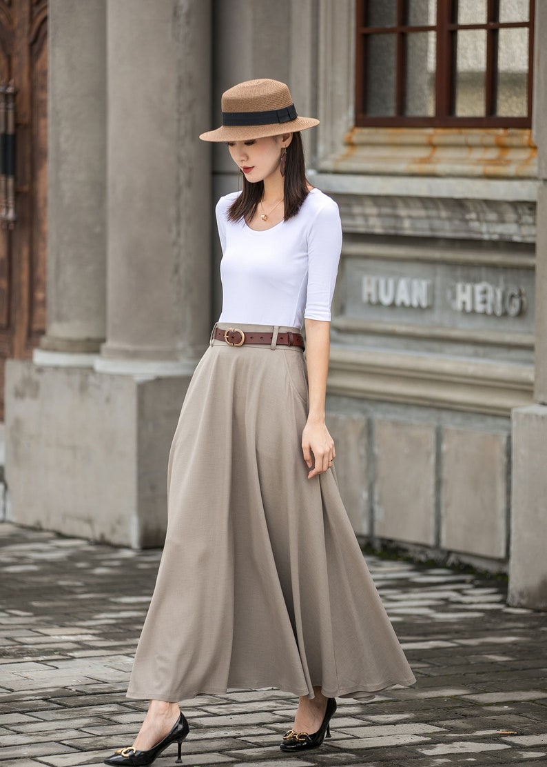 May include: A woman wearing a white top with elbow-length sleeves, a long beige skirt, and a brown belt. She is also wearing a brown hat with a black band and black high heels. The background features a building with the text "HUAN HENG".
