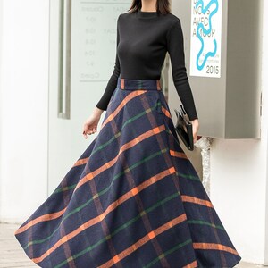 May include: A long, navy blue skirt with a plaid pattern in orange and green. The skirt is A-line and falls to the ankles. The model is wearing a black turtleneck sweater and a black hat.