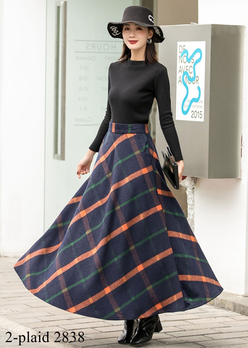 May include: A long, navy blue skirt with a plaid pattern in orange and green. The skirt is A-line and falls to the ankles. The model is wearing a black turtleneck sweater and a black hat.