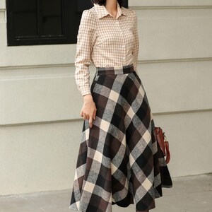 May include: A woman wearing a beige plaid button-down shirt and a long, flared plaid skirt in shades of brown, black, and cream. She is also wearing brown ankle boots. A brown leather handbag is visible.