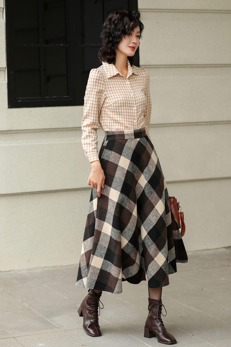 May include: A woman wearing a beige plaid button-down shirt and a long, flared plaid skirt in shades of brown, black, and cream. She is also wearing brown ankle boots. A brown leather handbag is visible.