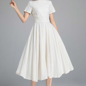 May include: A white linen dress with a boat neck, short sleeves, and a fitted bodice. The skirt is full and falls to the mid-calf.