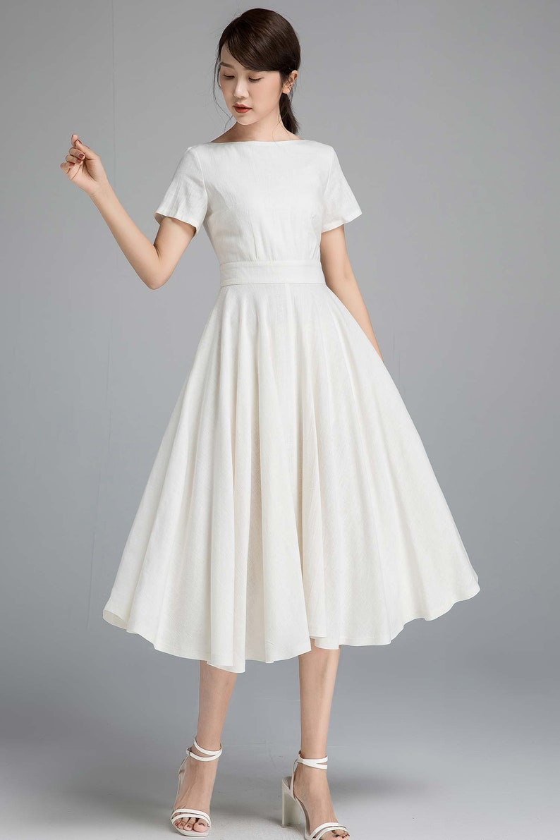 May include: A white linen dress with a boat neck, short sleeves, and a fitted bodice. The skirt is full and falls to the mid-calf.
