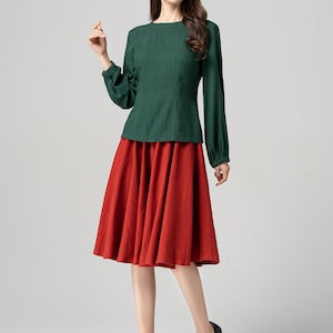 May include: A woman wearing a green long-sleeved blouse with a red midi skirt and black heels.