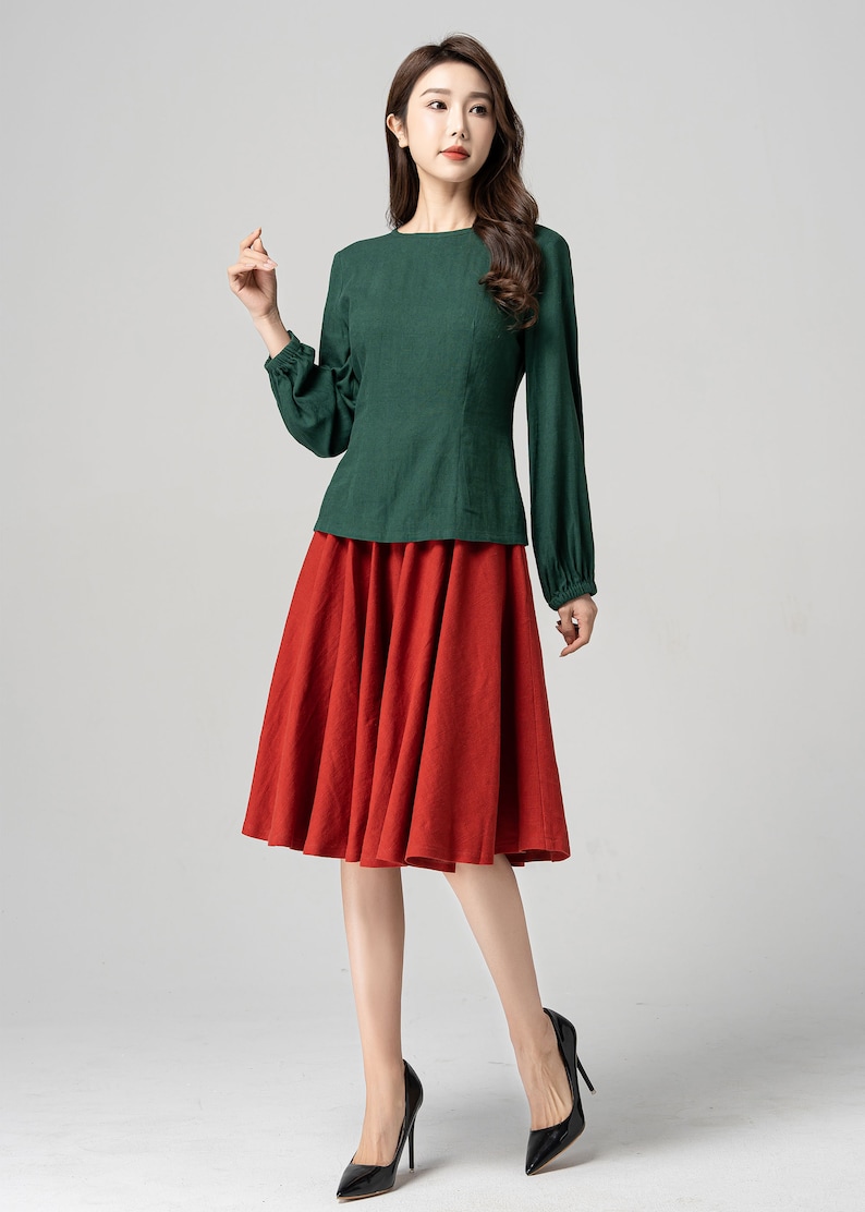 May include: A woman wearing a green long-sleeved blouse with a red midi skirt and black heels.