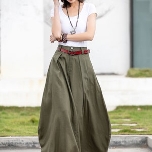 May include: A woman wearing a white t-shirt, a straw hat, and a long green skirt with a red belt. She is wearing sunglasses and a necklace.
