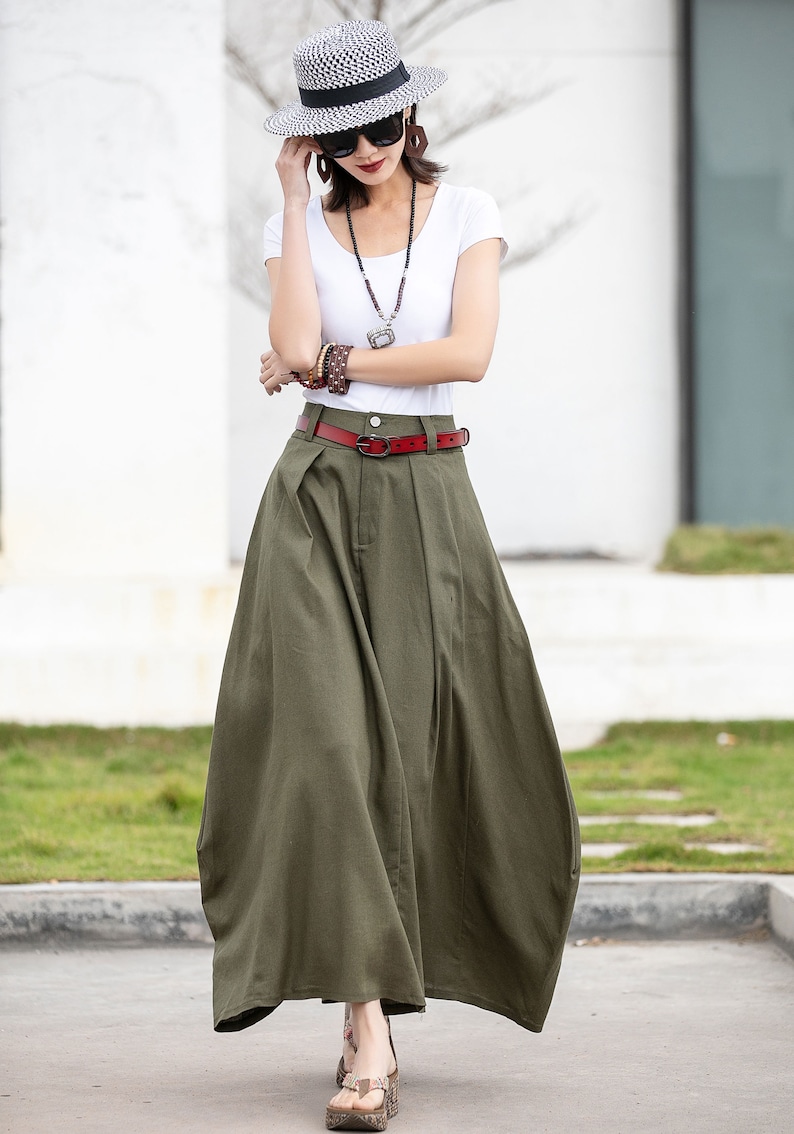May include: A woman wearing a white t-shirt, a straw hat, and a long green skirt with a red belt. She is wearing sunglasses and a necklace.