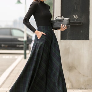 May include: A woman wearing a black turtleneck sweater and a black hat with a wide brim. She is wearing a long, green and black plaid skirt with a black purse. The skirt has pockets and a high waist.