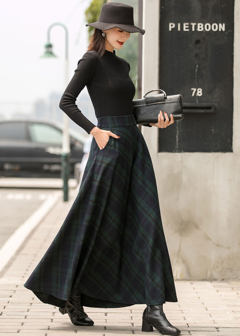 May include: A woman wearing a black turtleneck sweater and a black hat with a wide brim. She is wearing a long, green and black plaid skirt with a black purse. The skirt has pockets and a high waist.