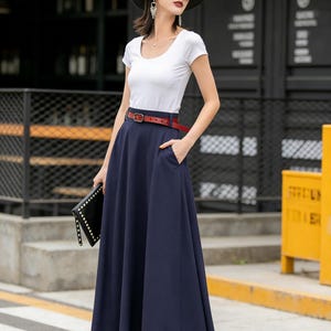 May include: A woman wearing a white scoop-neck t-shirt, a navy blue maxi skirt, and a red belt. She is also wearing a black hat, sunglasses, and holding a black clutch. The skirt has pockets.