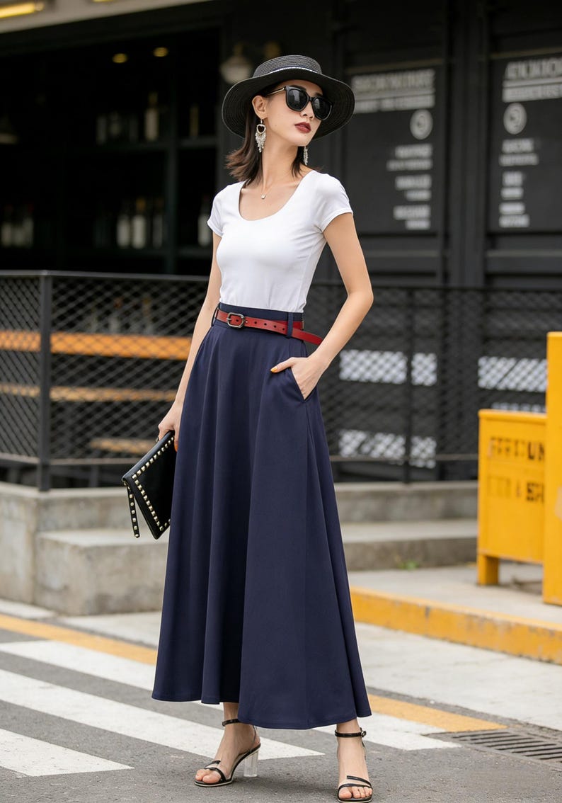 May include: A woman wearing a white scoop-neck t-shirt, a navy blue maxi skirt, and a red belt. She is also wearing a black hat, sunglasses, and holding a black clutch. The skirt has pockets.